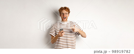 Technology and e-commerce concept. Amazed redhead man checking out online promo offer on mobile phone, pointing finger at smartphone and smiling, white background Technology and e-commerce concept. Amazed redhead man checking out online promo offer on mobile phone, pointing finger at smartphone and smiling, white background 98919782