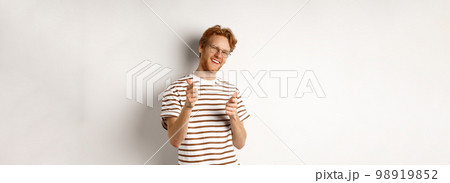 Cool and sassy redhead man with beard and glasses, pointing finger pistols at camera and smiling, congratulating you, standing over white background 98919852