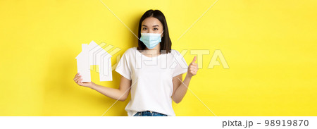 Covid-19, pandemic and real estate concept. Pretty asian woman smiling in medical mask, showing paper house cutout and thumb up, approve and like agency for buying property 98919870