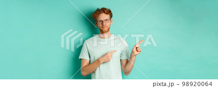Judgemental redhead man in glasses grimacing, frowning disappointed and pointing left, showing bad promo offer, standing over turquoise background 98920064