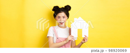 Real estate and family concept. Cute asian woman with bright makeup and stylish hairbuns, showing paper house cutout, smiling determined, yellow background 98920069