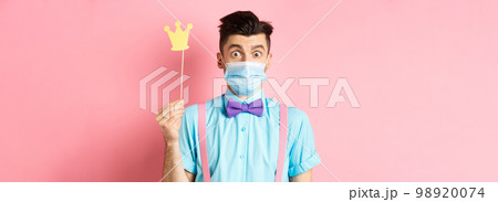 Covid, pandemic and quarantine concept. Cute and silly guy in medical mask looking surprised, holding small crown for party, standing over pink background Covid, pandemic and quarantine concept. Cute and silly guy in medical mask looking surprised, holding small crown for party, standing over pink background 98920074