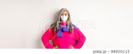 Covid, pandemic and social distancing concept. Confident and cheerful asian senior woman in respirator looking upbeat, smiling and holding hands on waist, white background 98920115