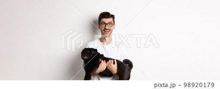 Happy hipster in glasses holding cute black pug and smiling, dog owner staring at camera with amazed smile, standing over white background 98920179