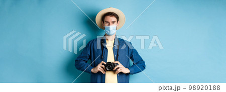 Covid-19, pandemic and travel concept. Young guy travelling abroad with camera, taking pictures on vacation, wearing medical mask from coronavirus, blue background 98920188