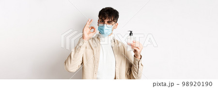Health, covid and quarantine concept. Portrait of natural guy in glasses and face mask showing bottle of good hand sanitizer, make okay sign, recommending product, white background 98920190