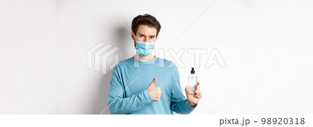 Covid-19, health and quarantine concept. Handsome smiling man in face mask, showing thumbs up and hand sanitizer bottle, recommending antiseptic brand, white background Covid-19, health and quarantine concept. Handsome smiling man in face mask, showing thumbs up and hand sanitizer bottle, recommending antiseptic brand, white background 98920318