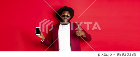 Handsome and stylish Black man showing phone screen and thumb up at camera, recommending online store app, red background Handsome and stylish Black man showing phone screen and thumb up at camera, recommending online store app, red background 98920339
