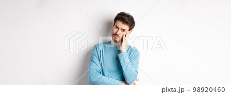 Sad young man looking frustrated, touching temple of head and frowning uneasy, standing against white background 98920460
