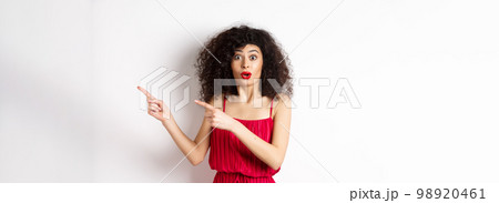 Surprised woman in red dress and makeup pointing fingers right, showing logo and look intrigued, standing on white background 98920461