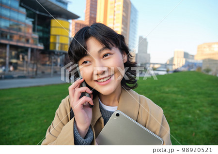 University lifestyle. Young asian student, girl with tablet and notebook, talks on mobile phone while walks from campus on street University lifestyle. Young asian student, girl with tablet and notebook, talks on mobile phone while walks from campus on street 98920521