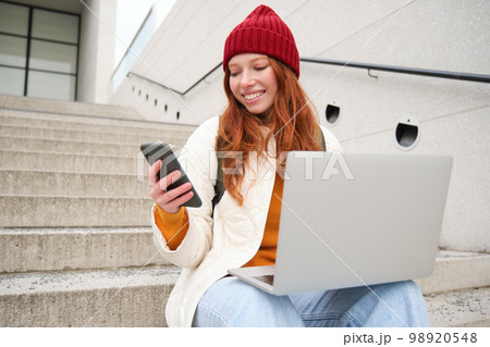 Young tourist, girl booking hotel room on laptop, enters confirmation code on smartphone, sitting with computer and phone on stairs outdoors 98920548