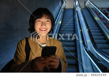 Young Japanese girl standing with smartphone, going down escalator with mobile phone, commuting in city, using public transport 98920549