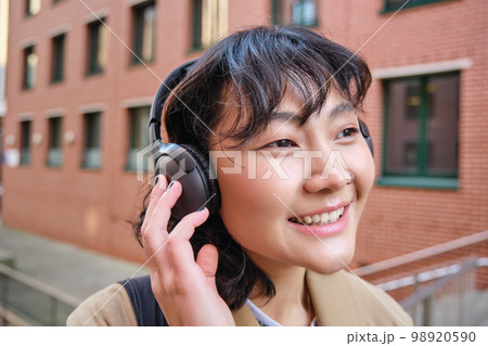 Close up portrait of stylish brunette asian girl, listens music in headphones, touches earphones and smiles, enjoys favorite song, stands on street Close up portrait of stylish brunette asian girl, listens music in headphones, touches earphones and smiles, enjoys favorite song, stands on street 98920590