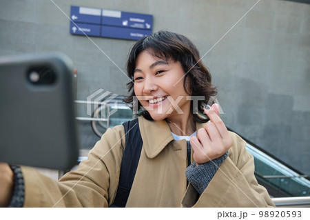 Portrait of stylish korean girl, student takes selfie on street, holds smartphone and poses on its camera, makes video on tourist trip, smiles with joy 98920593