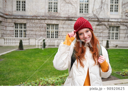 Beautiful redhead woman with backpack, tourist walking around city park and looks around, wears red hat and warm coat 98920625