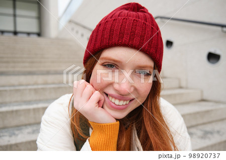 Beautiful redhead student, girl in red hat, smiles sincere, looks happy and relaxed, sits on stairs outdoors Beautiful redhead student, girl in red hat, smiles sincere, looks happy and relaxed, sits on stairs outdoors 98920737