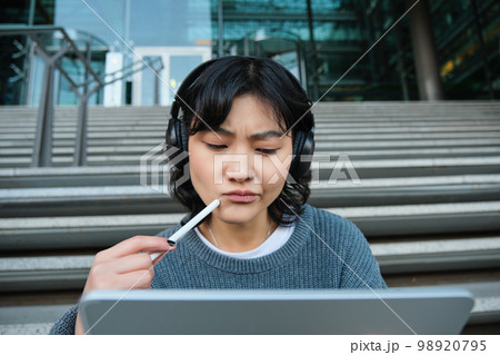 Portrait of asian girl with troubled face, draws on digital tablet with pen, looks complicated at her work, feels stuck no inspiration, sits on stairs and thinks Portrait of asian girl with troubled face, draws on digital tablet with pen, looks complicated at her work, feels stuck no inspiration, sits on stairs and thinks 98920795