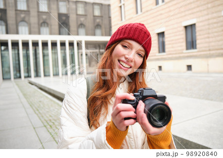 Beautiful readhead girl, photographer with professional camera takes pictures outdoors, walking around city and taking photos, sightseeing 98920840