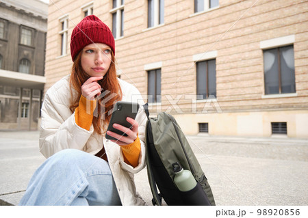 Young redhead woman with smartphone, sitting outdoors with backpack, thinking, looking thoughtful, making decision, looking at map and plans route Young redhead woman with smartphone, sitting outdoors with backpack, thinking, looking thoughtful, making decision, looking at map and plans route 98920856