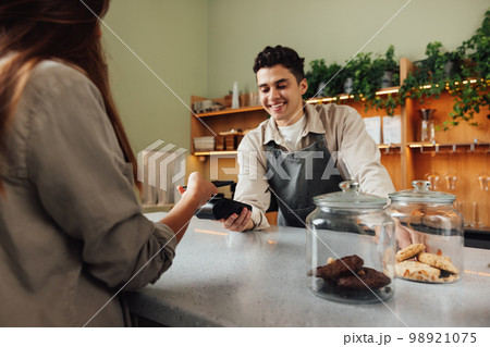 Smiling male barista holds a card machine while the customer pays by NFC. Barista in an apron receiving payment in a cafe at the counter. 98921075