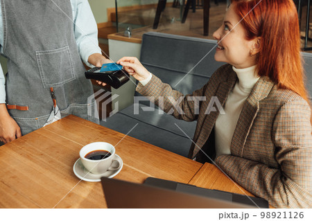 Woman paying by credit card with NFC in a coffeeshop. Female with ginger hair sitting at the table paying her bill. Woman paying by credit card with NFC in a coffeeshop. Female with ginger hair sitting at the table paying her bill. 98921126