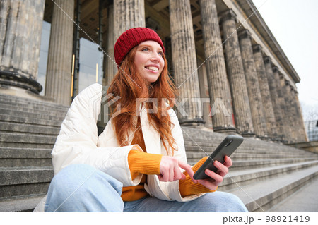 Portrait of young urban girl in red hat, sits on stairs near museum, holds mobile phone, connects to public wifi and surfs net, uses smartphone apps Portrait of young urban girl in red hat, sits on stairs near museum, holds mobile phone, connects to public wifi and surfs net, uses smartphone apps 98921419