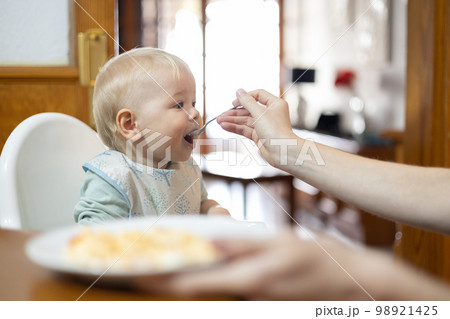 Mother spoon feeding her infant baby boy child sitting in high chair at the dining table in kitchen at home. 98921425