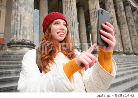 Portrait of young blogger, redhead girl tourist takes pictures of sightseeing, makes photos on smartphone camera, records video and smiles Portrait of young blogger, redhead girl tourist takes pictures of sightseeing, makes photos on smartphone camera, records video and smiles 98921442