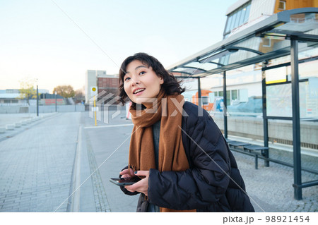 Cute young girl, student waiting on a bus stop, looking for her public transport on app tracker, holding smartphone, standing on street near road 98921454