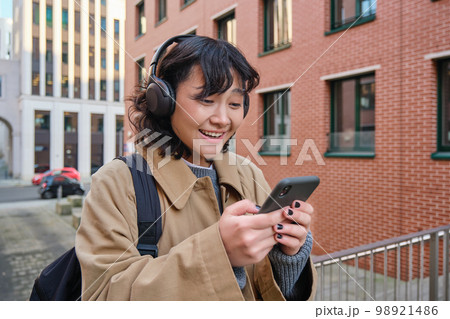 Happy korean girl looks at mobile phone and listens music in headphones, stands on street in city centre, reads text message on smartphone Happy korean girl looks at mobile phone and listens music in headphones, stands on street in city centre, reads text message on smartphone 98921486