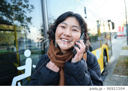 Cellular technology and people concept. Stylish asian girl talks on mobile phone, makes a telephone call, stands near bus stop and has conversation Cellular technology and people concept. Stylish asian girl talks on mobile phone, makes a telephone call, stands near bus stop and has conversation 98921504