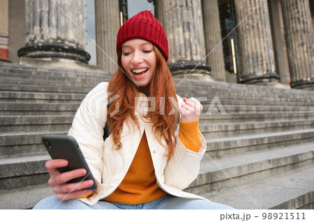 Achievement celebration. Happy redhead girl sits on stairs outdoors and looks at phone, triumphs, wins something and looks satisfied at mobile screen 98921511