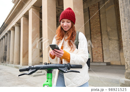 Beautiful young woman, tourist with backpack, rents e-scooter to travel around town, uses mobile phone application 98921525
