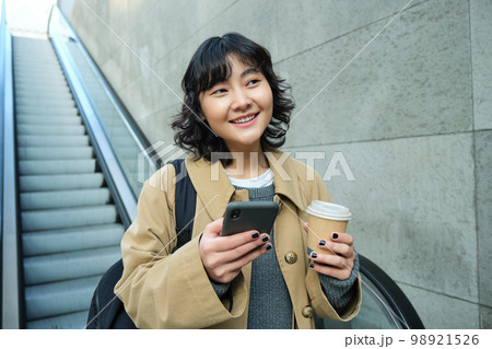Beautiful brunette girl drinks coffee to go, goes down escalator and smiles, holds smartphone, uses mobile app 98921526
