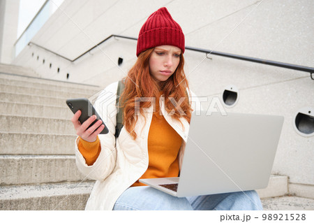 Portrait of redhead woman sits on stairs, uses laptop and holds smartphone, looks confused and upset at computer screen, tries to connect to wifi public internet 98921528