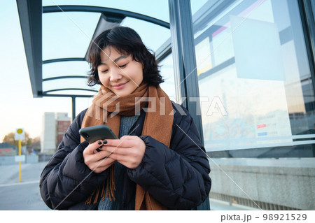 Beautiful Japanese girl, student standing on bus stop in winter jacket, holding smartphone, using application to track her public transport, ordering a ride 98921529