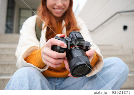 Smiling redhead girl photographer, checks her shots, holds camera and looks at screen, takes photos outdoors, walks around street and does streetstyle shooting 98921873