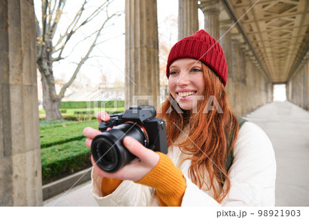 Young redhead female photographer, makes lifestyle shooting in city centre, takes photos and smiles, looks for perfect shot, makes picture 98921903