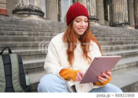 Outdoor shot of young stylish redhead girl sits on staircase and connects to public wifi, uses digital tablet, reads news on gadget Outdoor shot of young stylish redhead girl sits on staircase and connects to public wifi, uses digital tablet, reads news on gadget 98921908