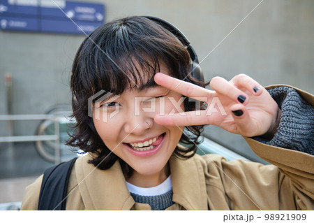 Head portrait of korean girl standing on street, showing peace sign, wearing headphones, listening music and smiling 98921909