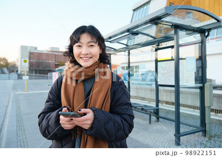 Happy smiling korean girl, using mobile phone, standing on bus stop with smartphone, looking at departure schedule on application, posing in winter clothes Happy smiling korean girl, using mobile phone, standing on bus stop with smartphone, looking at departure schedule on application, posing in winter clothes 98922151