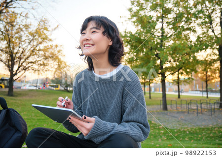 Young korean woman sits in park, draws on digital tablet, writes in gadget with pen tool, looks around and makes notes, creates artwork or scatches Young korean woman sits in park, draws on digital tablet, writes in gadget with pen tool, looks around and makes notes, creates artwork or scatches 98922153
