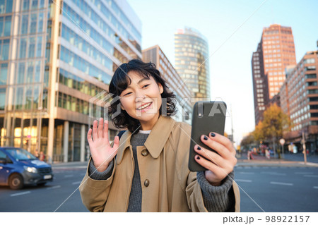 Happy asian girl tourist, takes selfie in city centre, makes video call and waves at smartphone camera, greets someone over phone Happy asian girl tourist, takes selfie in city centre, makes video call and waves at smartphone camera, greets someone over phone 98922157