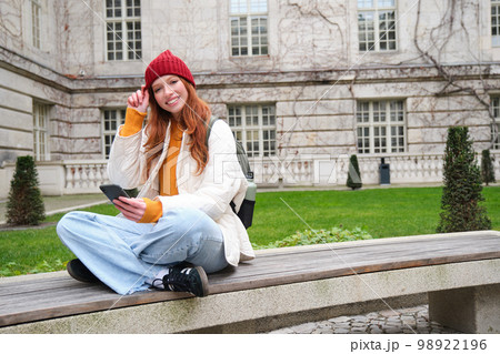 Young smiling redhead girl sits on bench and uses smartphone app, reads news online, watches video on mobile phone while relaxed in park 98922196