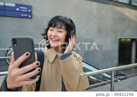 Beautiful korean girl in headphones, takes selfie on her smartphone, stands on street with mobile phone, records video, listens music and travels around city 98922298