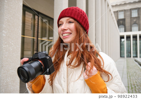 Smiling redhead girl photographer, taking pictures in city, makes photos outdoors on professional camera 98922433
