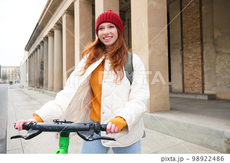 Smiling redhead european girl drives public e-scooter, tourist explores city, rides in city centre Smiling redhead european girl drives public e-scooter, tourist explores city, rides in city centre 98922486