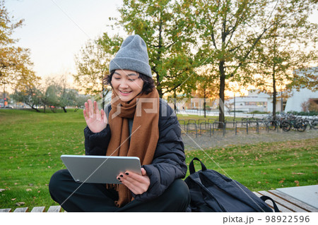 Portrait of young asian girl sits in warm clothes in park, waves hand at tablet, video chats outdoors, says hello while on call 98922596