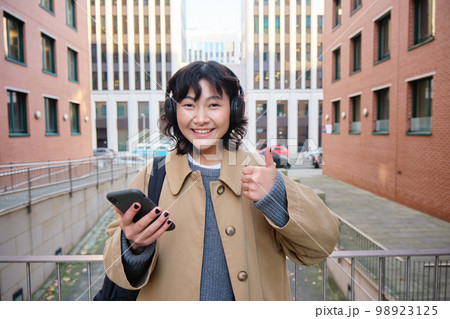 Portrait of cheerful asian girl in headphones, stands on street and shows thumbs up, found something online on her smartphone, using mobile phone Portrait of cheerful asian girl in headphones, stands on street and shows thumbs up, found something online on her smartphone, using mobile phone 98923125